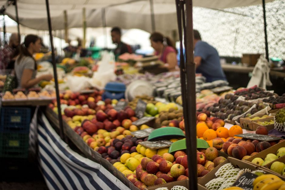 The image depicts a busy outdoor market stall under a canopy, with a long wooden table filled with a variety of fresh fruits neatly arranged in rows and piles. Visible fruits include apples in shades of red, yellow, and green, oranges, and peaches, all with smooth and slightly shiny surfaces reflecting natural light. The table is covered with striped fabric at the edges, and small blackboards or tags are placed among the produce for pricing or identification. Several people, some wearing casual clothing and gloves for handling produce, are seen behind the stall, slightly blurred in the background, engaged in arranging or selling the fruit. The environment suggests a bustling market atmosphere, with diffused natural light filtering through the canopy and possibly surrounding trees or open space behind the scene. The presentation of the fruit and the activity of vendors align with typical independent market or private vendor setups, akin to local rubbish removal and waste management practices associated with marketplace waste and spoilage, making this scene relevant in a context such as rubbish clearance or organic waste disposal services provided by companies like Rubbish Clearance Brixton.