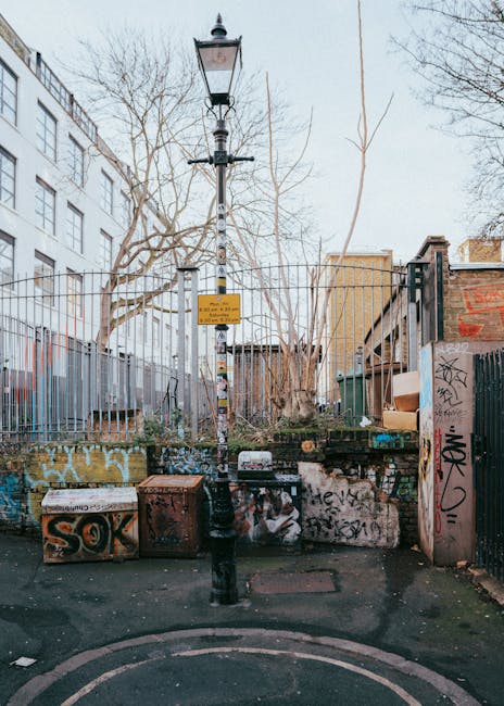 The image depicts an outdoor urban scene featuring a tall, black, vintage-style street lamp with a glass-enclosed lantern at the top, situated in the centre of a narrow paved area. The lamp post is marked with various stickers and remnants of graffiti, and is positioned in front of a low stone wall, which is also heavily covered in graffiti tags and painted with spray paint in multiple colours including orange, black, and white. Behind the wall, there is a fence made of vertical metal bars, painted in a dark colour, which runs along the left side of the image. The environment appears to be a back street or alleyway with a mix of residential and commercial elements, surrounded by a combination of brick and painted walls, some of which have additional graffiti. To the right, there is a small gate with graffiti markings. Leafless trees with bare branches extend into the sky above, indicating a winter or early spring setting. The ground is slightly worn with some debris and a metal manhole cover visible in the foreground, highlighting a neglected urban area typical of a location where private or alternative waste handling might be undertaken by services such as Rubbish Clearance Brixton. The overall scene is lit with natural daylight, giving it a neutral, everyday appearance.