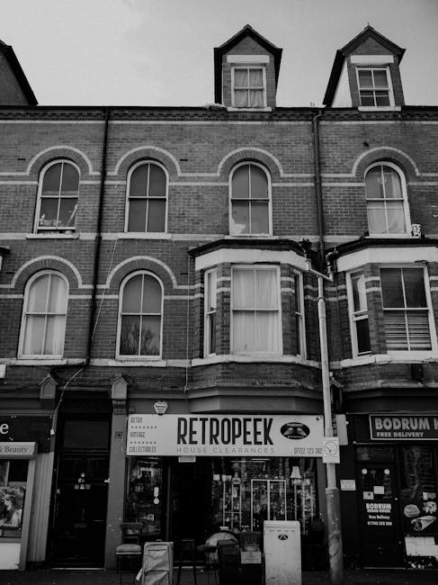 A three-storey brick building with a retail shop occupying the ground floor, prominently displaying a large sign that reads 'RETROPEEK' indicating house clearances. The upper floors feature four arched windows with white frames, three of which are visible along the facade, and the attic has two dormer windows with triangular gables, also with white-framed sash windows. The shopfront has glass windows and doors, with some items and signage visible inside, and is flanked by neighboring storefronts, including a fast food outlet with a sign that suggests free delivery. In front of the building, there are two benches and a small waste bin, indicating parking or pedestrian use. The background shows the upper part of the building under a cloudy sky, and the scene is captured in black and white, emphasizing the architectural details and urban environment. This image relates to alternative waste handling options through the visible house clearance business on-site.