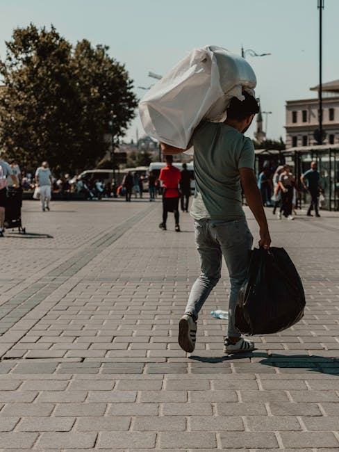A young man walking along a paved urban square during daytime, carrying a large white garbage bag slung over his shoulder and a black plastic bag in his right hand. The man is dressed in casual clothing, including a light green T-shirt, light grey ripped jeans, and sneakers. In the background, there are several other pedestrians, some walking and others standing, with a row of parked cars, trees, and buildings visible under a clear sky. The scene suggests a context of waste or rubbish collection, possibly indicating private disposal or preparatory work for rubbish removal services, with the setting being a public space where individual waste handling is taking place.