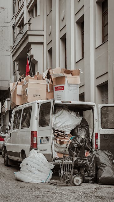 A white cargo van parked on a city street in front of a multi-story building with a grey facade and several windows. The van's rear doors are open, revealing an interior filled with black garbage bags, cardboard boxes, and various discarded materials stacked inside and spilling onto the pavement. Multiple large cardboard boxes, some labeled with red and black printing, are placed on top of the van, with additional boxes and waste bags positioned on the ground nearby. A small hand trolley loaded with black rubbish bags and broken-down cardboard is situated against the rear of the van. The scene captures a typical urban rubbish removal setup, illustrating independent waste collection or on-site clearance that Rubbish Clearance Brixton might facilitate, set against the backdrop of the building's exterior environment and street pavement. The overall atmosphere is neutral and professional, with natural lighting highlighting the textures of the waste and the urban surroundings.