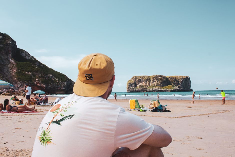 A person wearing a beige cap with a black patch that reads 'BRIXTON' and a white t-shirt featuring a tropical bird and plant design, sitting on a sandy beach with their back facing the camera. The beach scene includes several sunbathers lying on towels and sitting under umbrellas, with some people walking along the shoreline. In the background, the ocean has gentle waves, with a large rocky island covered in greenery rising from the water. The sky is partly cloudy with soft, natural lighting illuminating the scene. The environment appears relaxed and summery, with leisure activities visible on the sand and in the sea. This setting highlights a typical seaside location suited for outdoor relaxation, and may relate to independent rubbish clearance or waste management services in outdoor or recreational areas managed by companies like Rubbish Clearance Brixton.