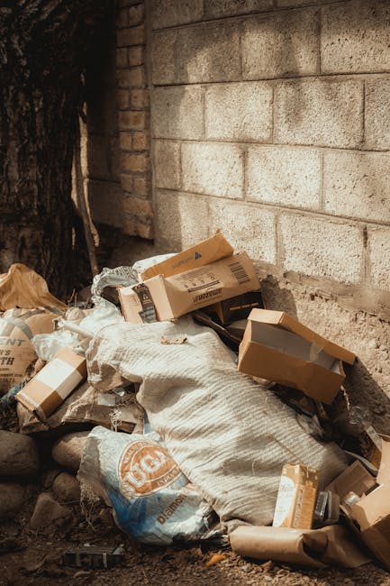 A pile of discarded waste and packaging materials, including flattened cardboard boxes, paper bags, and a large woven sack, rests on uneven ground next to a rough brick wall and the base of a tree. The cardboard boxes vary in size and have labels and printing on their surfaces, with some partially flattened or crumpled. The woven sack appears to contain additional rubbish, with its fabric showing signs of wear. The ground is composed of dirt and small rocks, and the background features a brick wall constructed from large, rectangular, light-colored bricks with visible mortar joints. To the left, part of a tree trunk with textured, dark bark is visible, providing natural contrast to the urban surroundings. The scene is illuminated by natural daylight, emphasizing the textures of the materials and the earthy tones of the environment. This image visually represents an area where private waste accumulation is awaiting collection, highlighting the type of refuse managed by independent rubbish clearance services such as Rubbish Clearance Brixton, which offers alternative waste handling to local authority disposal.