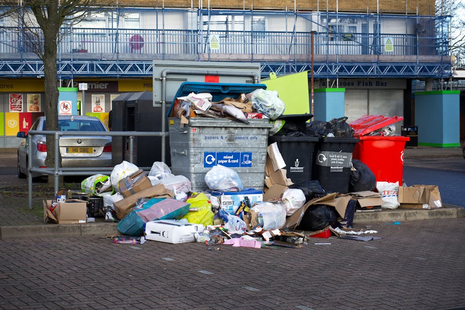 A busy street scene featuring a variety of household and commercial rubbish laid out for collection or disposal. In the foreground, several items are visible, including a large, dark-colored sofa with a textured fabric finish, positioned on the pavement near the curb. Adjacent to the sofa, multiple smaller objects such as a lamp with a fabric shade, a small table, and various scattered household items are seen. Some objects are stacked or leaned against each other, creating an irregular pile of waste situated directly outside shops and service providers. Behind the rubbish, there are storefronts with signs indicating a pharmacy, a fish and seafood shop, and other retail outlets, with the shop fronts and windows occupying the middle ground. People are gathered around the waste collection area, some engaging in conversation, others inspecting the items, or walking past. The backdrop includes a multi-story brick building with large windows, and a tree on the left side partially visible, casting shadows onto the scene. The lighting appears natural, suggesting daytime with diffuse sunlight. This image illustrates a typical scene where private waste clearance services, such as those offered by Rubbish Clearance Brixton, manage street-level rubbish collection of bulky items, complementing the area's waste disposal options.